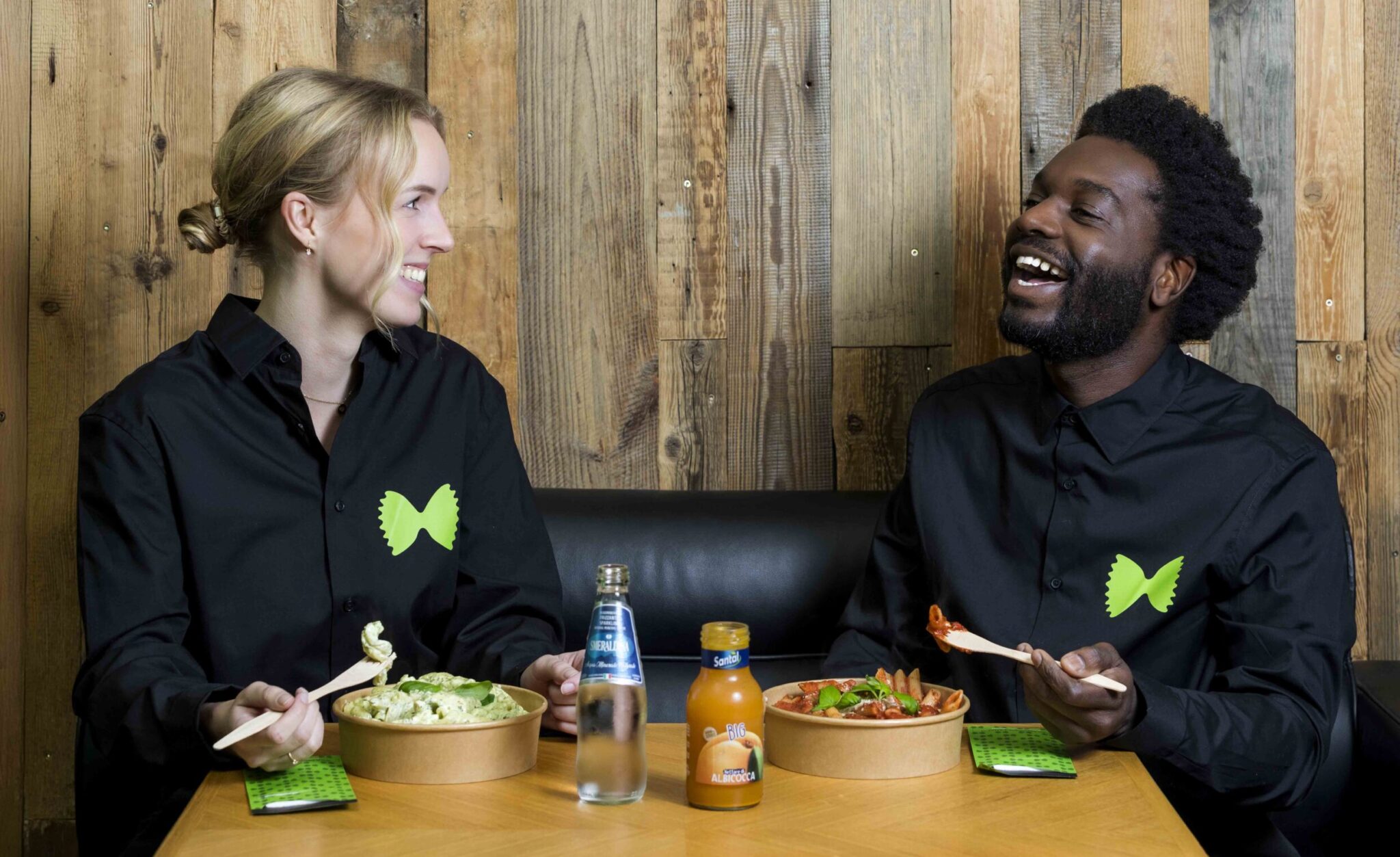 Twee medewerkers van Helpman's Pasta's in zwarte shirts lachen terwijl ze samen verse pasta eten